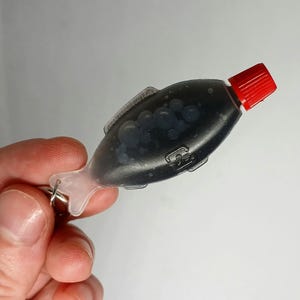 Pu&ograve; includere: Un portachiavi a forma di pesce che rappresenta una bottiglia di salsa di soia con tappo rosso. Il liquido scuro &egrave; visibile attraverso la plastica trasparente. Il portachiavi &egrave; attaccato a un anello d'argento.