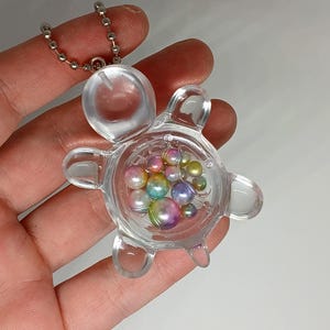 May include: Clear plastic turtle-shaped keychain filled with iridescent, colorful beads. The keychain has a silver ball chain attached to the top. The beads inside are various sizes and colors, including pink, yellow, and purple.