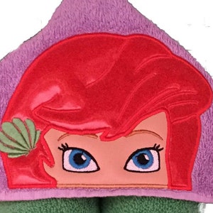 May include: A hooded towel featuring an embroidered design of a cartoon character's face with red hair, blue eyes, and a green seashell. The towel is purple and green, suitable for bath time.