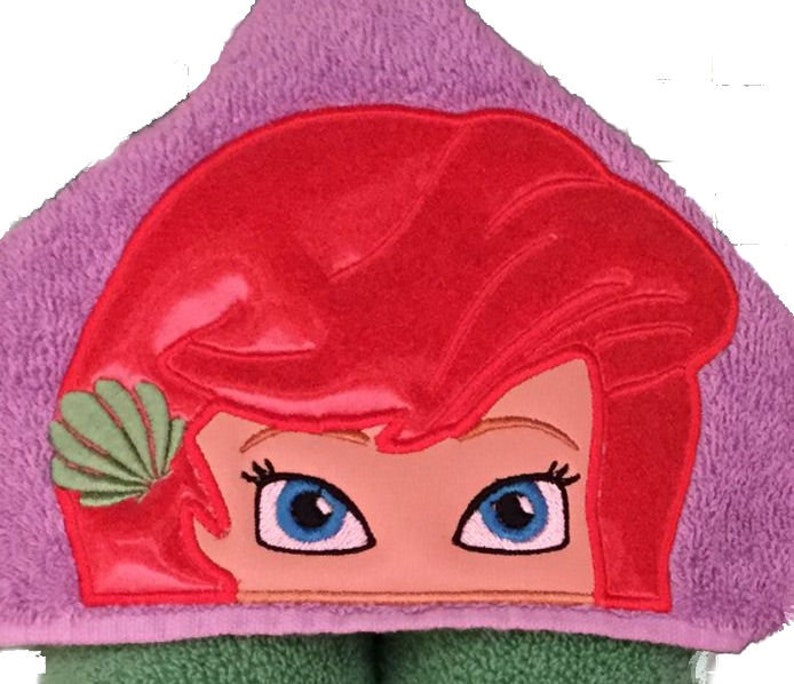 May include: A hooded towel featuring an embroidered design of a cartoon character's face with red hair, blue eyes, and a green seashell. The towel is purple and green, suitable for bath time.