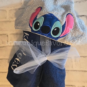 May include: Blue hooded towel with a stitched-on cartoon character of Stitch from Lilo & Stitch. The towel is tied with a white tulle ribbon and the word "Izzy" is embroidered on the towel.