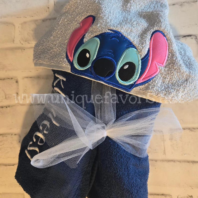 May include: Blue hooded towel with a stitched-on cartoon character of Stitch from Lilo & Stitch. The towel is tied with a white tulle ribbon and the word "Izzy" is embroidered on the towel.