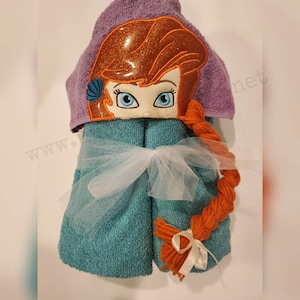 May include: A teal hooded towel featuring an embroidered design of a cartoon character's face with orange hair and blue eyes. The hood is purple. The towel is tied with a white ribbon and tulle.