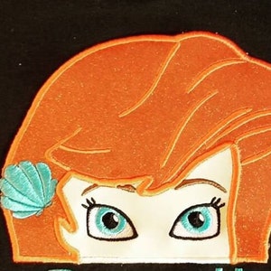 May include: Embroidered patch featuring a cartoon character's face with orange hair and blue eyes. A turquoise seashell adorns the hair. The character's face is outlined in black, with a white background.