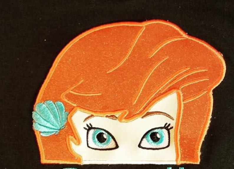 May include: Embroidered patch featuring a cartoon character's face with orange hair and blue eyes. A turquoise seashell adorns the hair. The character's face is outlined in black, with a white background.