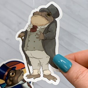 May include: A sticker of a cartoon frog wearing a top hat, vest, and coat. The frog is holding a cane and has a serious expression on its face.