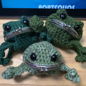 May include: Three crocheted frog coin purses in shades of green. Each frog has a silver coin purse opening with a decorative clasp.
