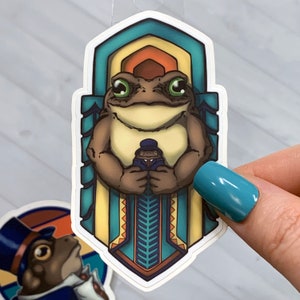 May include: A sticker featuring a cartoon toad wearing a top hat and holding a smaller toad in a suit. The toad is standing in a decorative archway with blue, green, and yellow accents.