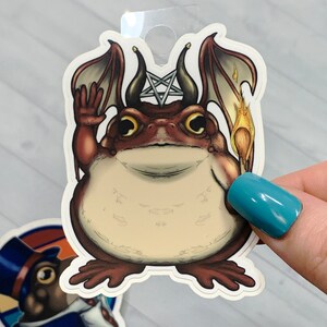 May include: A sticker featuring a cartoon toad with wings, a pentagram, and a flaming torch. The toad is wearing a crown and has a mischievous expression.