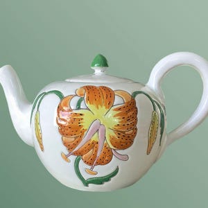 Op de afbeelding: Witte keramische theepot met een groot ontwerp van een oranje en gele leliebloem. De theepot heeft een gebogen handvat, een tuit en een groene knop op het deksel. Groene en gele accenten maken het bloemmotief compleet.