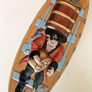 Può includere: Un piatto decorativo ovale in ceramica con una scena dipinta di un pirata in cappotto rosso e cappello nero con emblema di teschio. Il pirata tiene un sacco di monete d'oro accanto a una botte di legno, un pappagallo e una corda sul ponte.