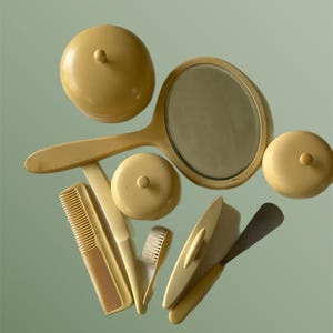 Puede incluir: Un conjunto de accesorios de tocador de color marfil vintage, que incluyen un espejo de mano, dos tarros con tapa, un peine, un cepillo y un calzador. Los artículos están dispuestos sobre una superficie verde claro, proyectando sombras suaves.
