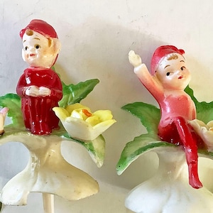 May include: Two vintage fairy figurines. Each fairy is wearing a red outfit and hat, and is perched on a white flower with green leaves and yellow flowers. One fairy is standing, the other is seated with an arm raised.