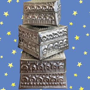 Puede incluir: Una pila de cuatro cajas de metal plateado con intrincados diseños en relieve. Las cajas están apiladas una encima de la otra, con la caja más grande en la parte inferior y la caja más pequeña en la parte superior.