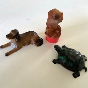 May include: Three animal figurines: a brown dog lying down, a brown dog standing on a red bottle cap, and a dark green geometric turtle. The dog figurines have blue eyes.