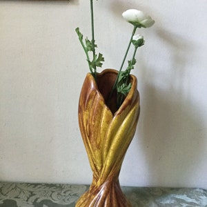 May include: A tall, ceramic vase in shades of brown and yellow, with a textured, leaf-like design. The vase holds three white artificial flowers with green stems and leaves. The vase is a decorative home accent.