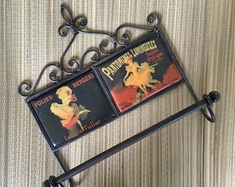 May include: A black metal towel rack with two decorative tiles. The tiles feature vintage illustrations of dancers and the text "Folies Bergeres" and "Pantomimes Lumineuses Theatre Optique L'Etrangle".