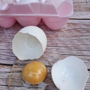 May include: A pink plastic egg carton containing several white eggs, with one broken egg in the foreground showing a clear, gelatinous egg white and a round, golden-brown yolk. The text Xohappycrafts.com is printed in the bottom right corner.