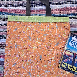 Peut inclure: Un sac fourre-tout avec des anses en tissu noir et une bordure supérieure vert clair à motif de squelettes. Le corps principal est orange avec un imprimé répétitif de squelettes, de sucettes et de bonbons. Un livre City of the Dead est visible.