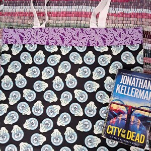 Peut inclure: Un sac fourre-tout noir avec des anses blanches arborant un motif répétitif de boules de cristal bleu clair tenues par des mains blanches. La bordure supérieure est violette avec un imprimé de chauves-souris et de lunes noires. Un livre est présent.