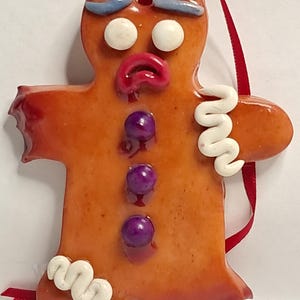 May include: A gingerbread man ornament with a red ribbon for hanging. The ornament has white icing details, purple candy buttons, and a red mouth. The gingerbread man has blue icing eyebrows and white candy eyes. The ornament is made of a glossy material.