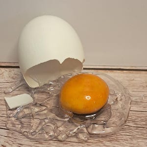 May include: A decorative sculpture of a cracked egg featuring a white shell fragment and a realistic, glossy orange yolk resting on a clear, textured resin base that mimics spilled egg white. The text Xohappycrafts.com is visible at the bottom.