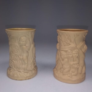 May include: Two beige ceramic vases with a raised design of cherubs and trees. The vases are about 4 inches tall.