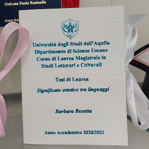 Può includere: Una carta bianca con testo turchese e un emblema turchese. La carta è legata con un nastro bianco e un nastro rosa. Il testo include il nome di un'università, un dipartimento, un corso e il titolo "Tesi di Laurea".