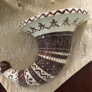 May include: A white ceramic cornucopia wall decor with brown and white geometric patterns. The cornucopia is shaped like a horn and has a wide opening at the top.