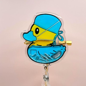 Op de afbeelding: Een doorzichtige, eendvormige badge reel met een gele eend die een blauwe chirurgische muts en masker draagt. De eend heeft zwarte ogen en houdt chirurgische instrumenten vast. De badge reel is bevestigd aan een goudkleurige metalen clip.