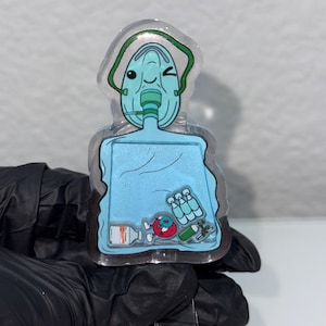 May include: A clear plastic badge holder featuring a cartoon oxygen mask with a winking face. The mask is connected to a blue bag containing a red donut, a bottle of hand sanitiser, and a green syringe. The badge holder has a clear plastic clip.