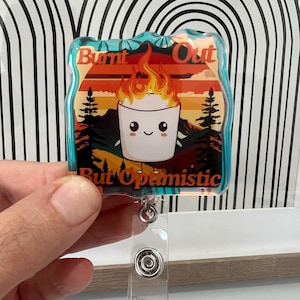 May include: A clear badge holder with a colourful graphic of a smiling marshmallow on fire with the text "Burnt Out But Optimistic".