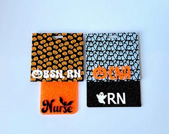 Pode incluir: Dois porta-crachás com tema de Halloween. Um apresenta um fundo preto com abóboras laranja e o texto "BSN, RN". O outro tem um fundo preto com fantasmas brancos e o texto "CNA, RN". Ambos têm detalhes em laranja.