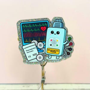 May include: A glittery badge reel featuring a cartoon-style medical theme. It includes a heart monitor, a cute battery-shaped character with the text "REPLACE BATTERY PLEASE", and a package of "Disposable ECG Electrodes".