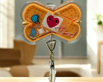 May include: A badge reel in the shape of a bandage, featuring a heart with a smiling face, medical equipment, and a face mask. The reel is orange with a clear plastic overlay and a metal clip.