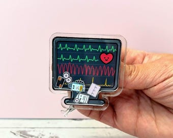 May include: A clear acrylic badge featuring a computer monitor displaying a heart rate monitor with green and red lines and a red heart. The badge also includes medical equipment icons. The badge is held in a hand.