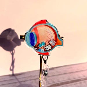 May include: Anatomy-themed badge reel featuring a detailed illustration of an eye cross-section. The design includes a blue iris, red and orange internal structures, and a small eye chart. The badge reel is attached to a gold-colored metal stand.