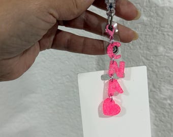May include: A white retractable badge holder with a pink heart and the letters CNA in pink glitter. The badge holder is attached to a white card.