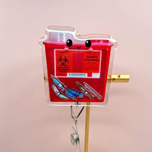 May include: A red and clear badge reel featuring a cartoon sharps container with a smiling face. The container has the words "SHARPS CONTAINER" and "BIOHAZARD INFECTIOUS WASTE" printed on it. The reel also has images of syringes and needles.