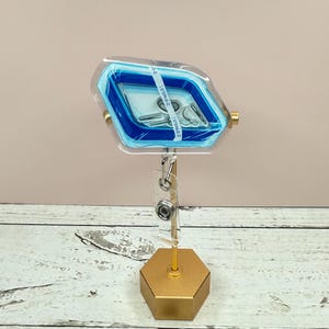 May include: A clear blue and white plastic badge holder with a gold metal stand. The badge holder has a blue border and a clear window for displaying a badge or ID card. The stand is hexagonal shaped and has a gold finish.