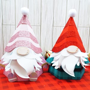 May include: Two papercraft gnomes, one with a pink and white striped hat and the other with a red and white striped hat. Both gnomes have white beards and are sitting on a red and white checkered surface.