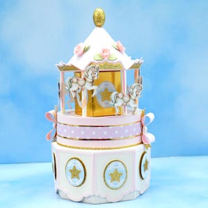 May include: A decorative carousel-themed box with a white and gold roof, pink and white polka dot and star-adorned tiers, and miniature carousel horses. The box features gold accents, pink bows, and a gold finial. The overall design is whimsical and elegant.