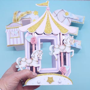 May include: A pink and gold carousel-themed papercraft with two white horses and a pink flower detail. The carousel is decorated with glitter and stars.