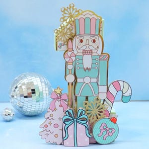 May include: A pastel-colored Christmas-themed display featuring a nutcracker, Christmas tree, gift, ornament, and candy cane. The nutcracker is teal, pink, and gold. A disco ball and small sphere are in the background. The tree, gift, and ornament are pink and teal.