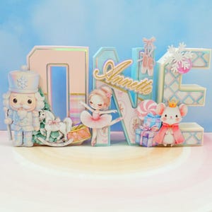 May include: A decorative "ONE" sign with a ballerina theme. The sign features a nutcracker, a ballerina, a mouse with a crown, and the name "Annette". The letters are pastel colors with glitter accents, and include a rocking horse and gifts.