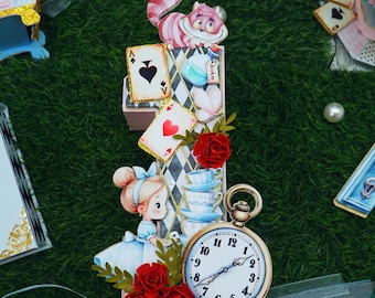 May include: A whimsical Alice in Wonderland-themed decorative piece. It features a Cheshire Cat, playing cards, a pocket watch, and a young girl in a blue dress. Red roses and teacups add to the fairytale aesthetic. The pocket watch shows the time.