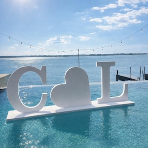 May include: White floating sculpture in a pool spelling out "C", a heart, and "L". The letters and heart are large and stand out against the blue water and sky. A string of lights hangs above the water.