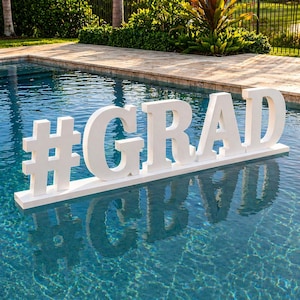 May include: A white, floating sign in a pool spells out #GRAD in large, block letters. The sign is reflected in the clear, blue water. The background includes greenery and a pool deck, suggesting a graduation celebration.