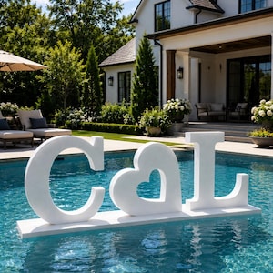 May include: A large white freestanding sign featuring the letters C and L separated by a heart shape, mounted on a rectangular base and floating on the surface of a swimming pool. The letters are solid white with a smooth, matte finish.