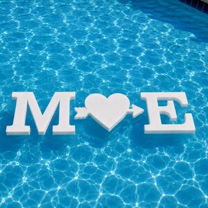 May include: A white floating decoration featuring the letter M, a heart shape pierced by an arrow, and the letter E. The letters and heart are constructed from a solid white material and are positioned horizontally on the surface of blue water.